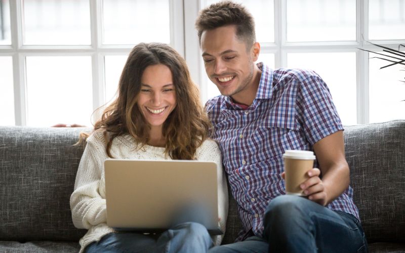 proofreading-services-happy-couple-laughing-looking-on-laptop-screen-sitting-on-sofa-min